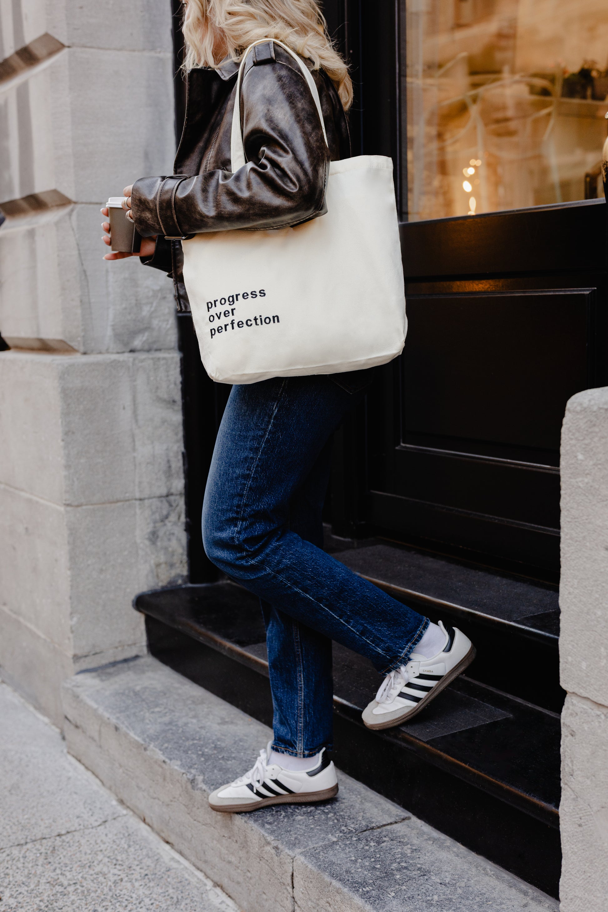 Person walking up steps holding tote bag with text Progress over Perfection, wearing jeans and sneakers.