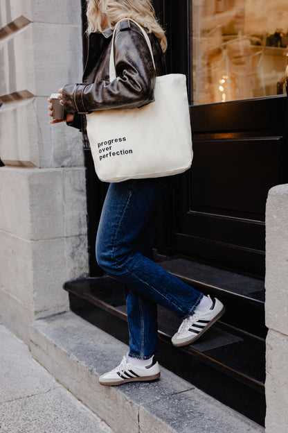 Person walking up steps holding tote bag with text Progress over Perfection, wearing jeans and sneakers.
