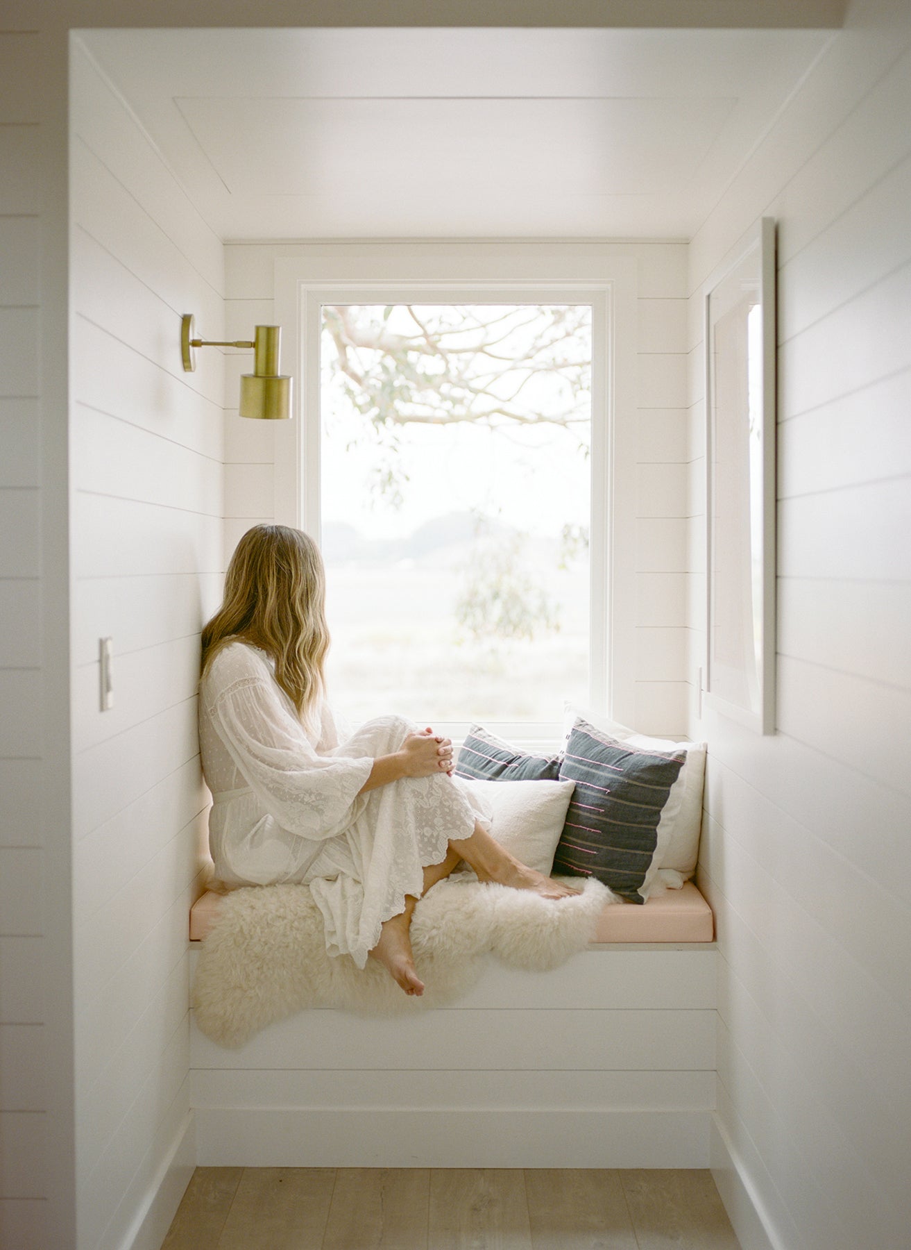 Woman sitting on a window seat with cushions in a bright room.