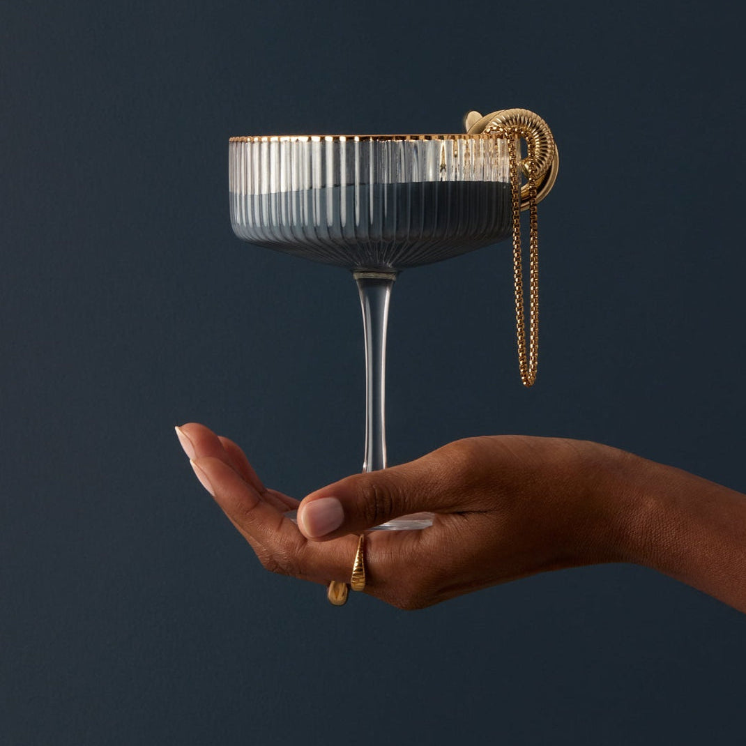 Glass held by hand against After Hours interior paint dark blue wall by Blu Paint with gold chain accessory