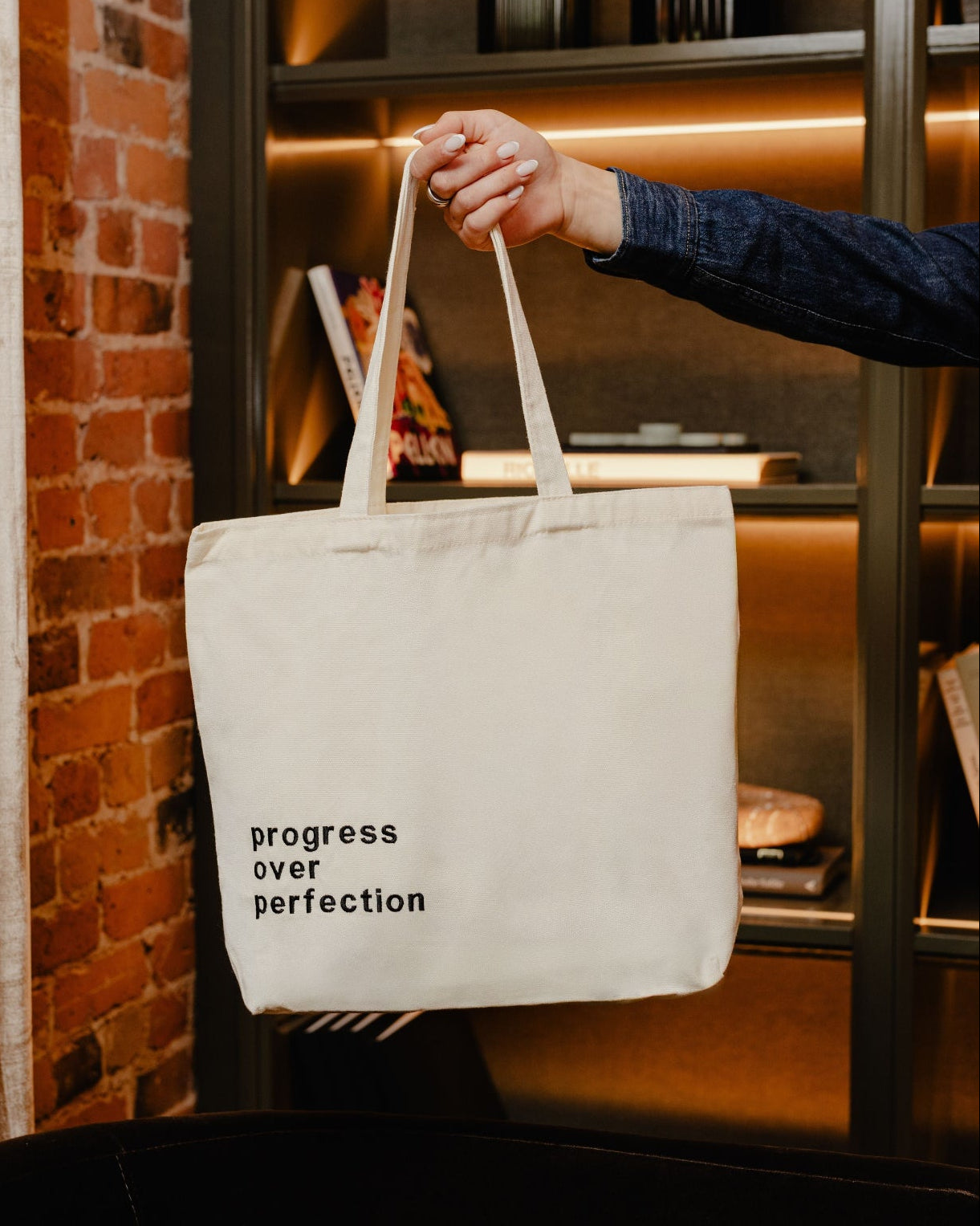 Tote bag with 'progress over perfection' text held by a person in an indoor setting.