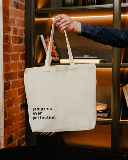 Tote bag with 'progress over perfection' text held by a person in an indoor setting.